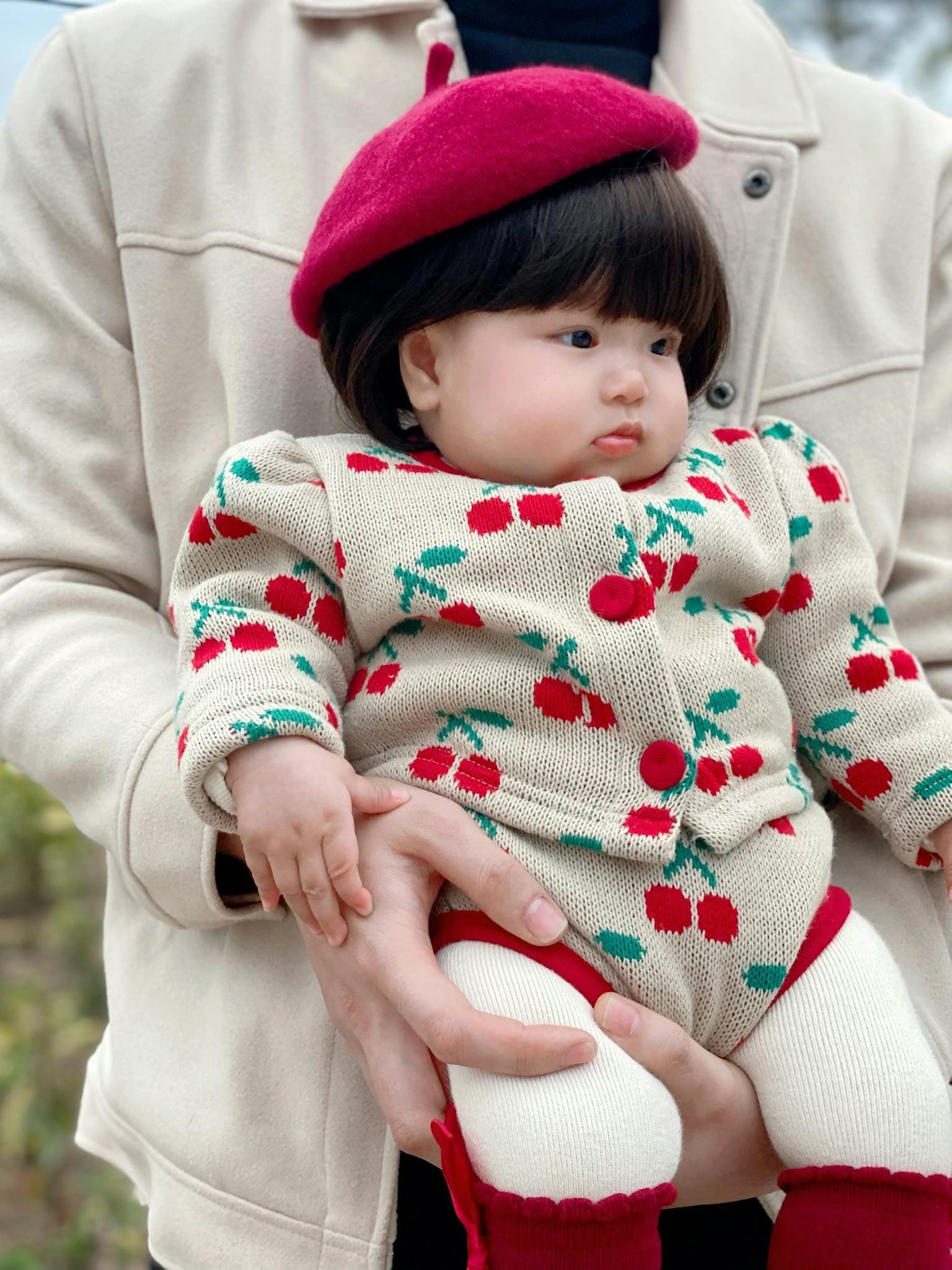 Adorable baby wearing cherry-themed knitwear and red beret held by an adult.