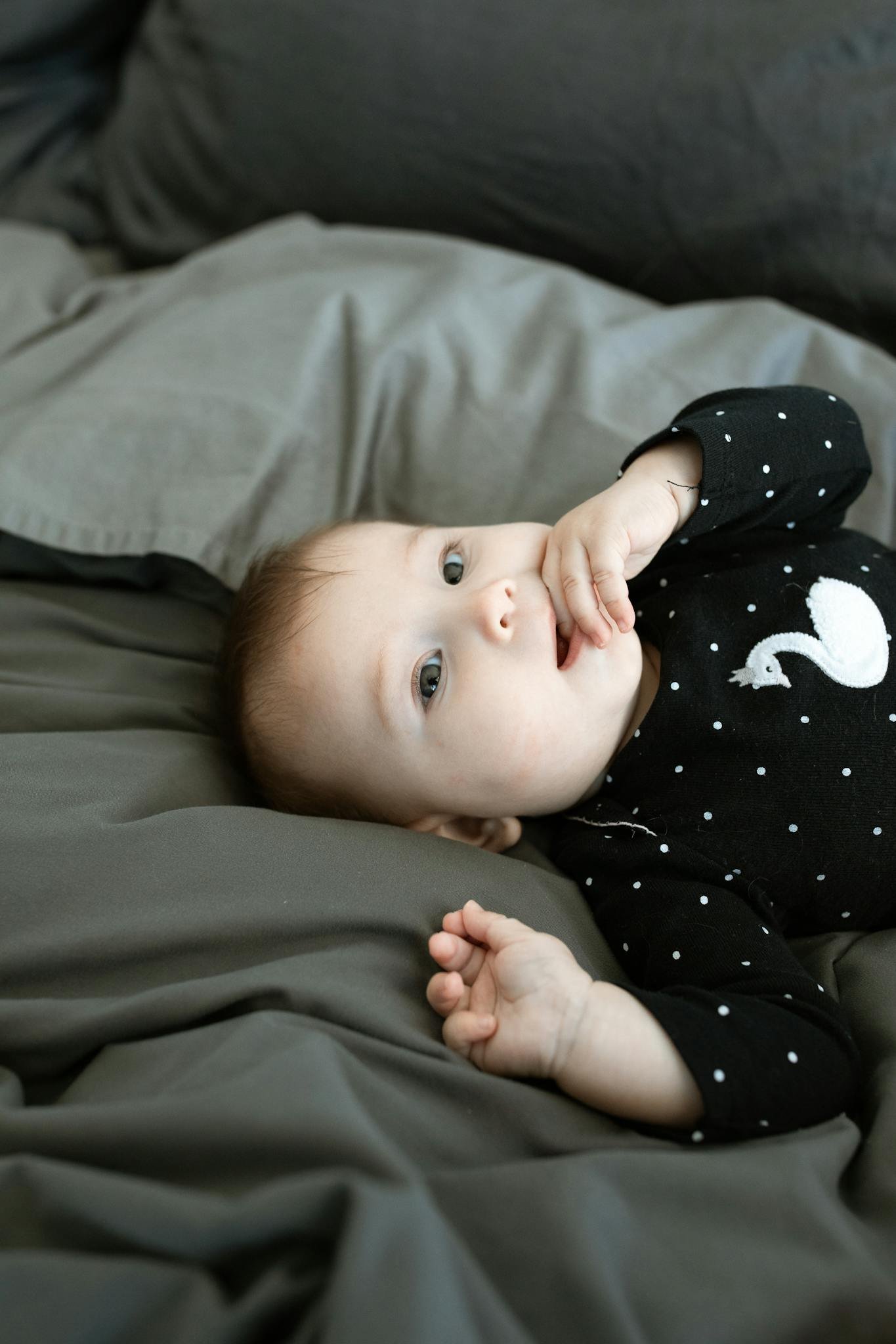 Cute baby girl in a black polka dot outfit lying on bed, sucking fingers.