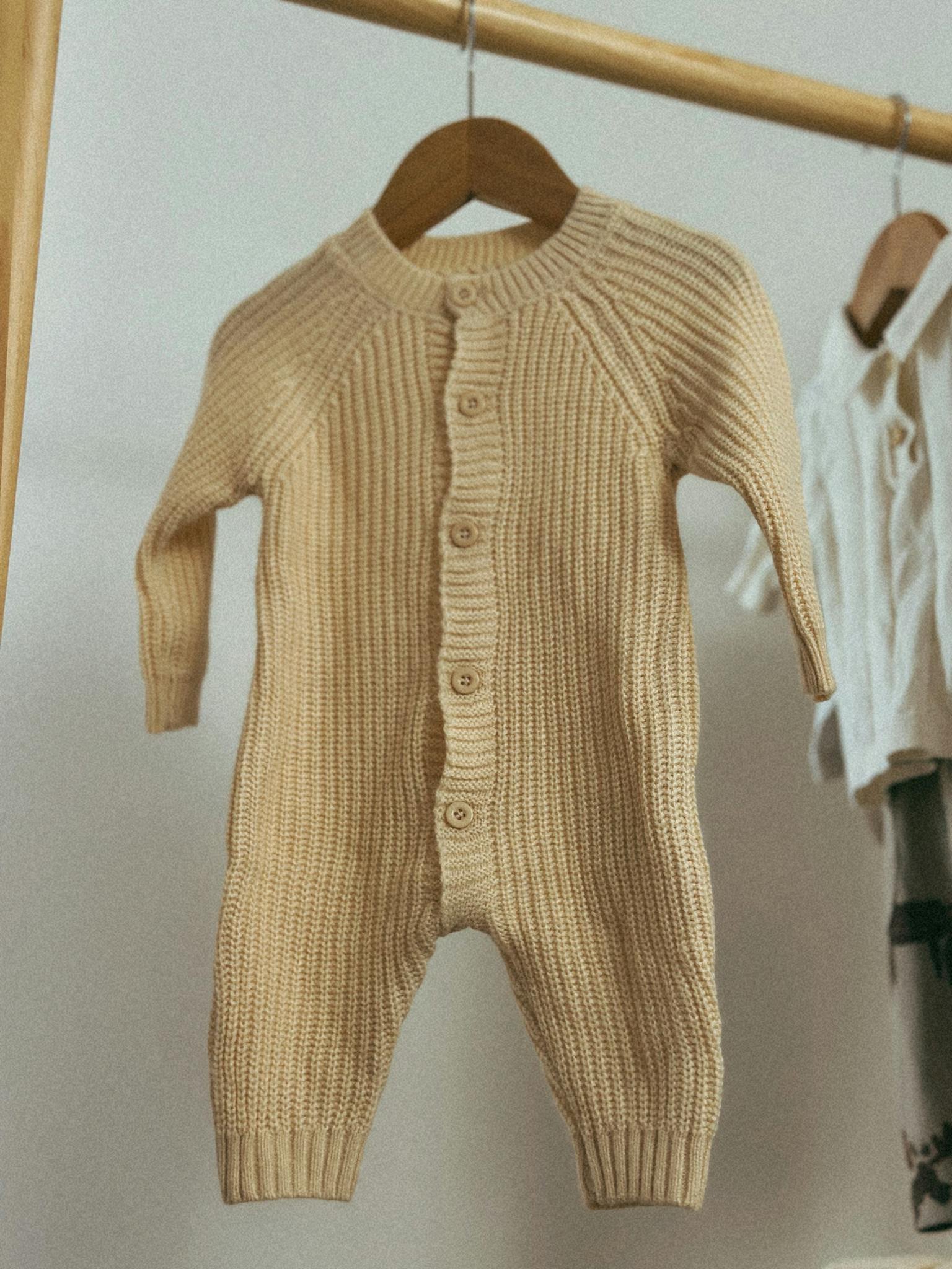 Warm knitted baby onesie in neutral tones hanging indoors on a wooden rack.
